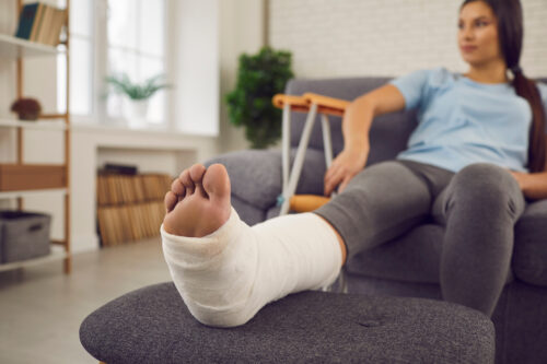 Concept of rehabilitation of people after serious physical accident injury. Female patient with broken leg in plaster cast sitting on sofa. Young woman with foot bone fracture resting on couch at home