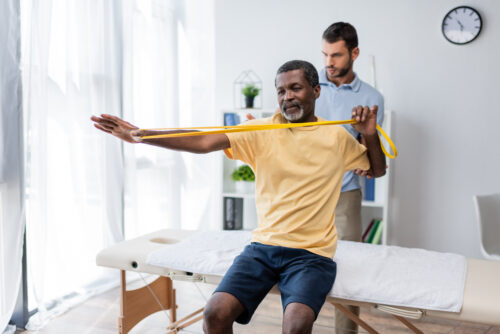 middle aged african american man sitting on massage table and training with elastics near physical therapy