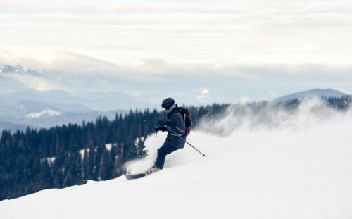 Panoramic photo with skier, winter mountains view. Young man skiing downhill from snow-capped mountain top. Risk, overcoming obstacles, hardiness, victory, achievement concept. Grey sky on background.